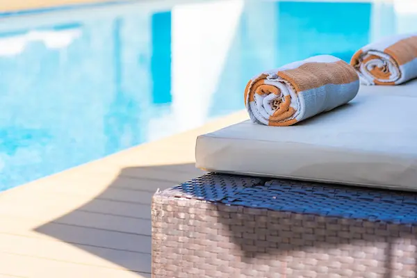 End of a pool chair with a rolled up towel on the bed with a hotel pool in the background